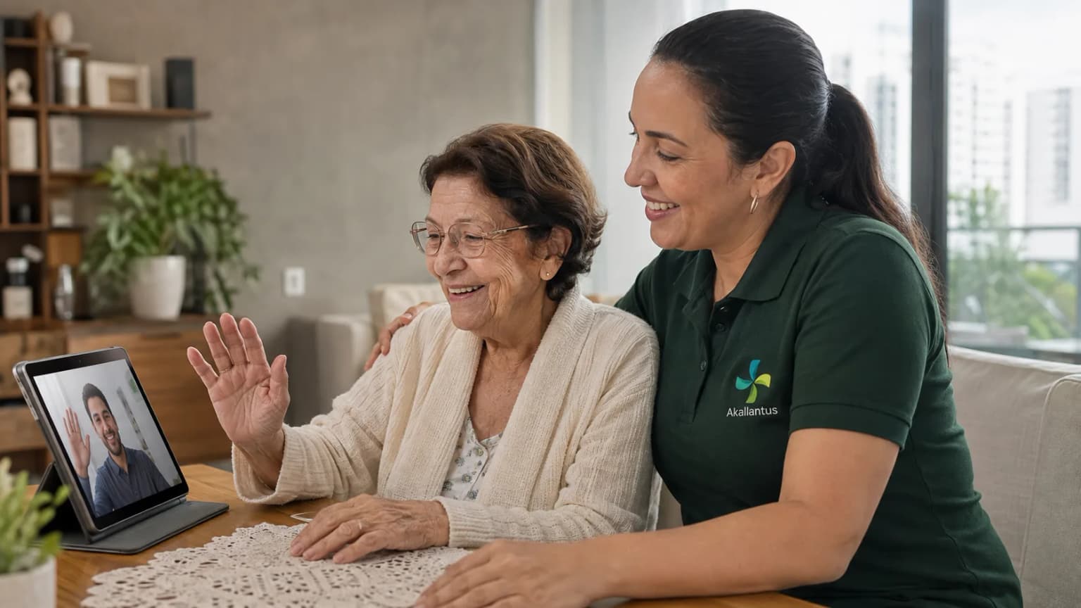 A caregiver in a green Akallantus polo sits beside an elderly woman during a video call with her son on a tablet, in a bright São Paulo living room.