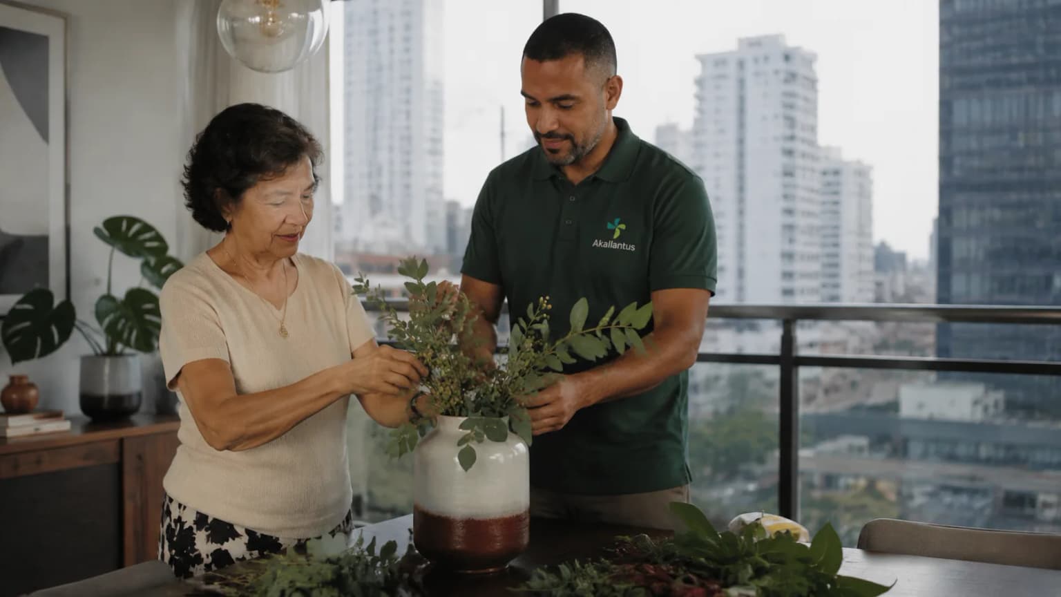 Cuidador da Akallantus arruma um arranjo de folhagens ao lado de uma senhora idosa em um apartamento com vista da cidade, no Centro Expandido de São Paulo.