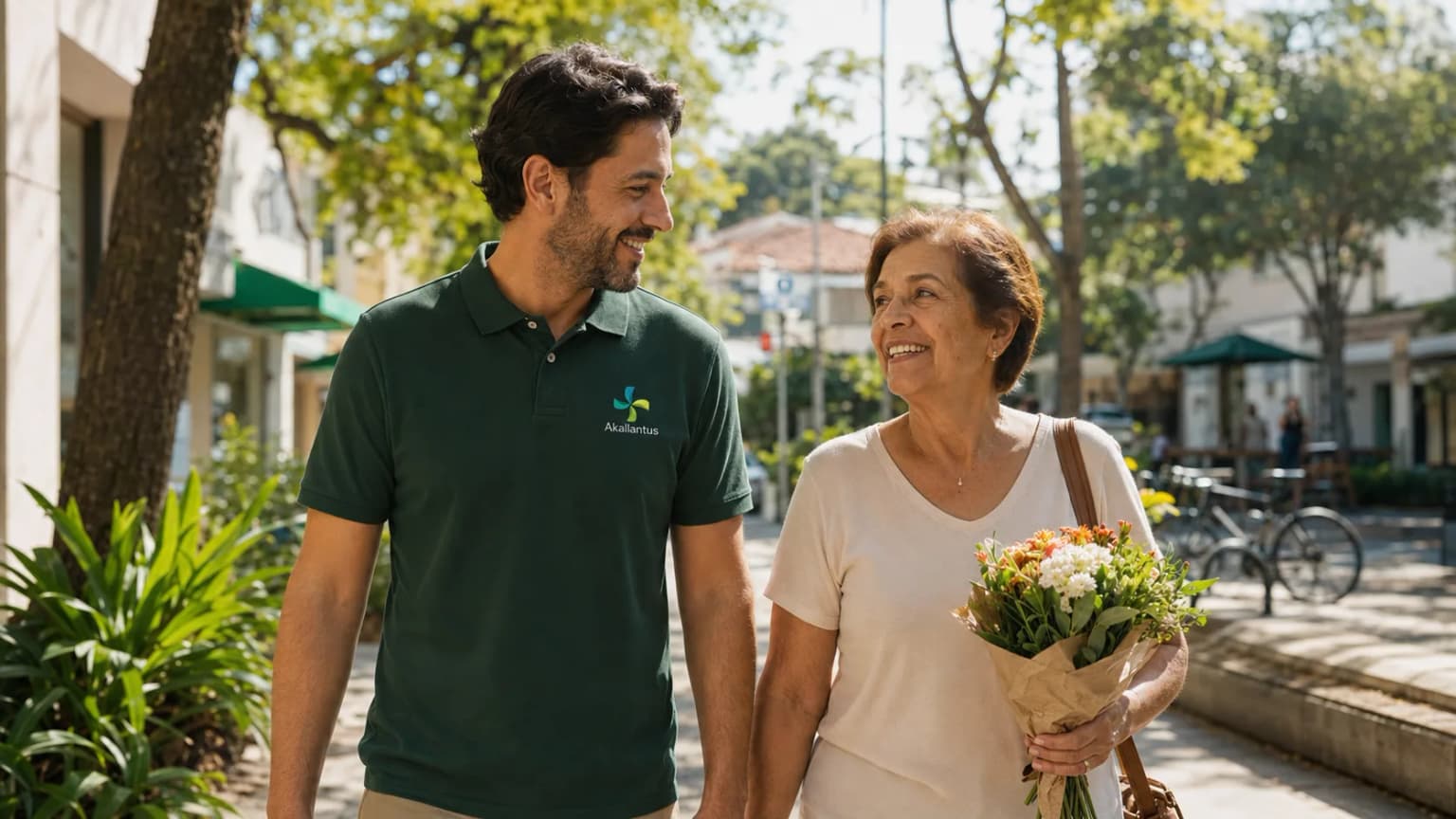 Cuidador da Akallantus caminha ao lado de uma senhora idosa que carrega um buquê de flores por uma rua arborizada em Moema, São Paulo.