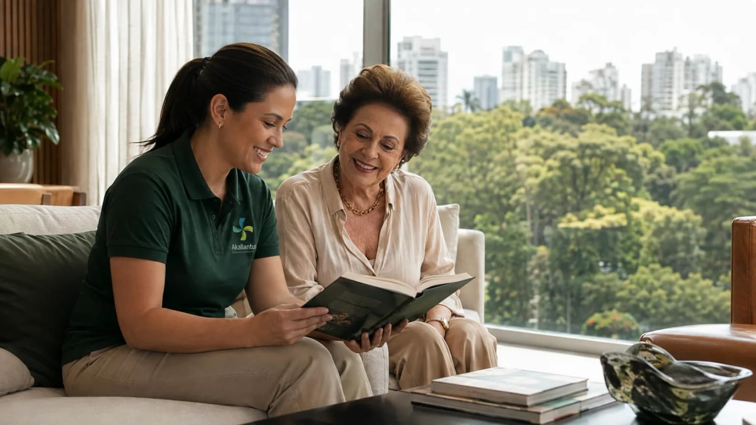 Cuidadora da Akallantus, de uniforme verde, lê um livro ao lado de uma senhora idosa em um apartamento de alto padrão no Panamby, São Paulo, com vista arborizada da cidade.