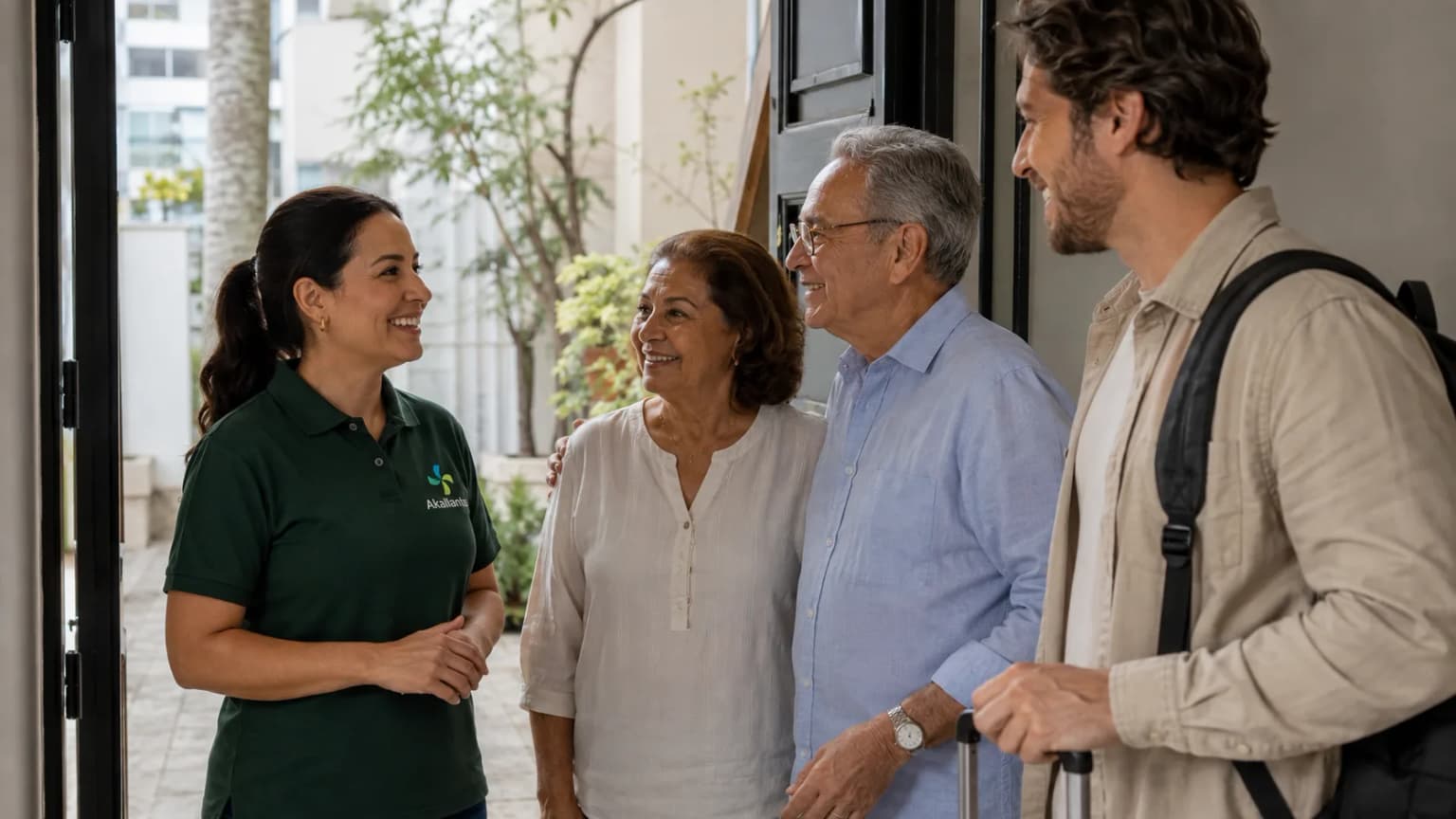 A caregiver in a green Akallantus polo welcomes an elderly couple and their adult son at the door of a São Paulo home, as the family arrives from abroad.