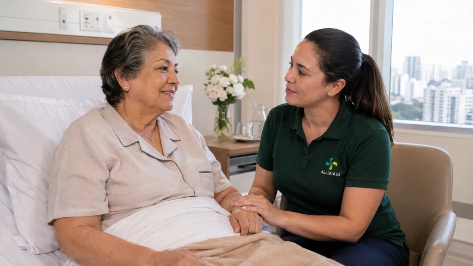 A caregiver in a green Akallantus polo holds the hand of an elderly woman resting in a hospital bed, offering calm companionship.